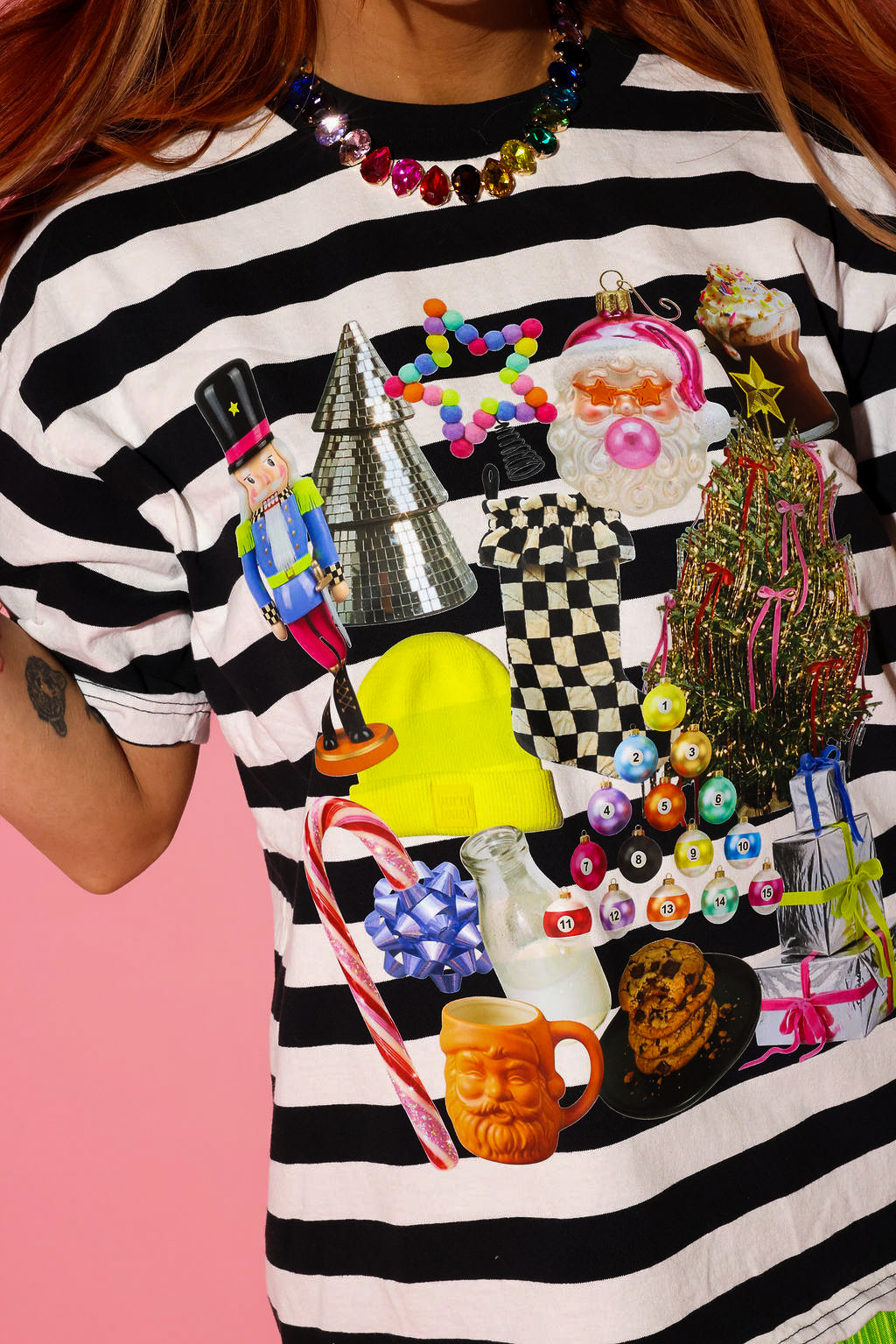 Model wearing a black and white striped tee with a Christmas graphic and a rainbow jeweled necklace on a pink background