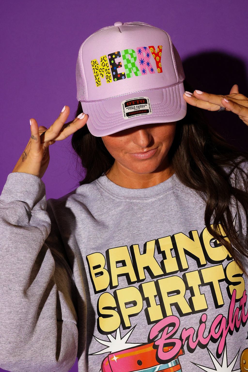 A model tilts a lavender trucker hat reading “MERRY” in multicolor patterned block letters down over her face, styled with a heather gray “Baking Spirits Bright” graphic crewneck sweatshirt. Background is purple.