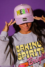 A model tilts a lavender trucker hat reading “MERRY” in multicolor patterned block letters down over her face, styled with a heather gray “Baking Spirits Bright” graphic crewneck sweatshirt. Background is purple.