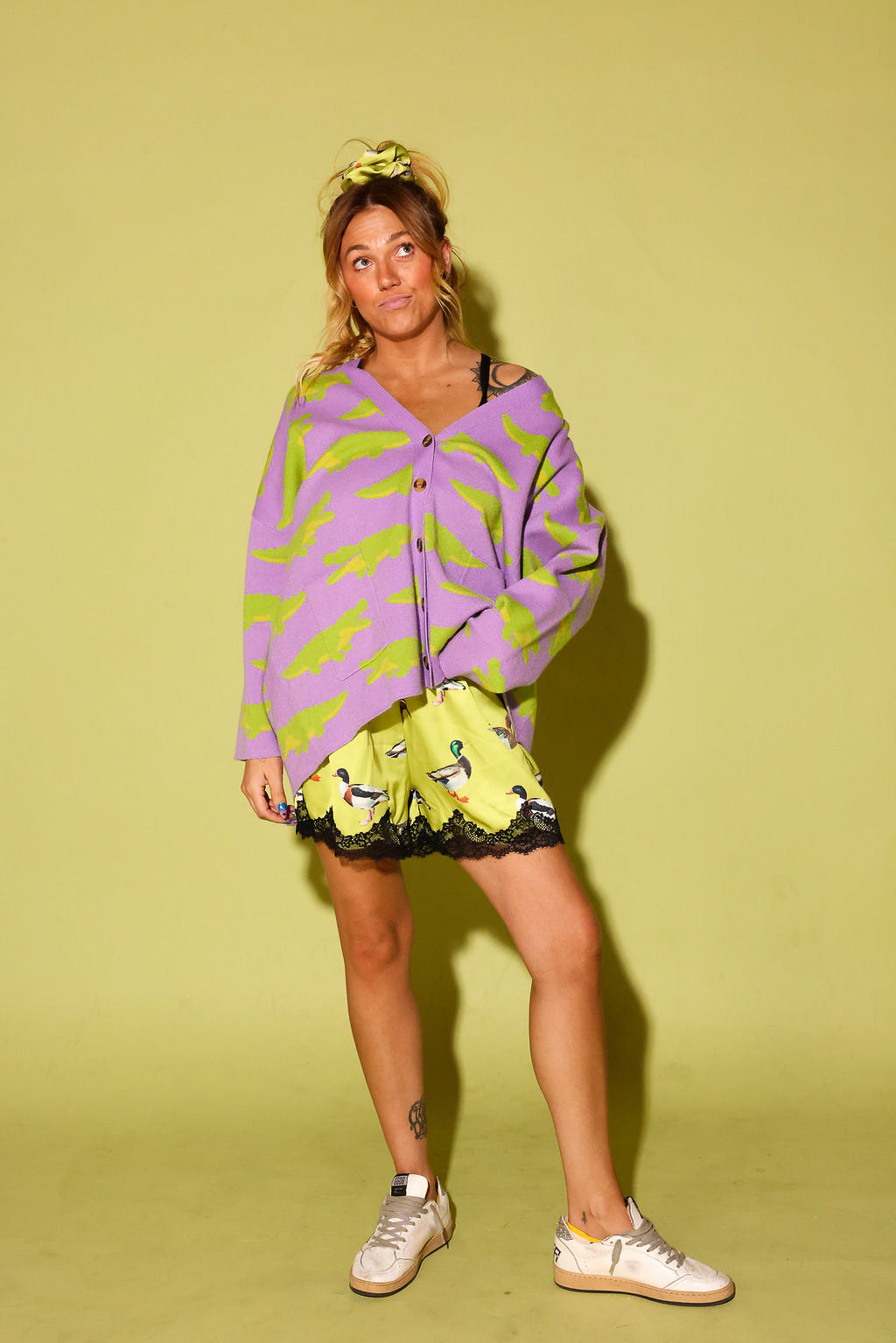 model wearing a purple cardigan with green alligators, and green satin shorts with black lace trim and a design, and white sneakers in front of a pale yellow background