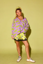 model wearing a purple cardigan with green alligators, and green satin shorts with black lace trim and a design, and white sneakers in front of a pale yellow background