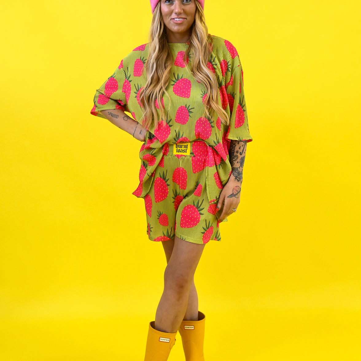 A woman modeling a bright gold and pink strawberry print set with a yellow burnt toast tag on waistband and pink beanie, paired with yellow rain boots, against a solid yellow background