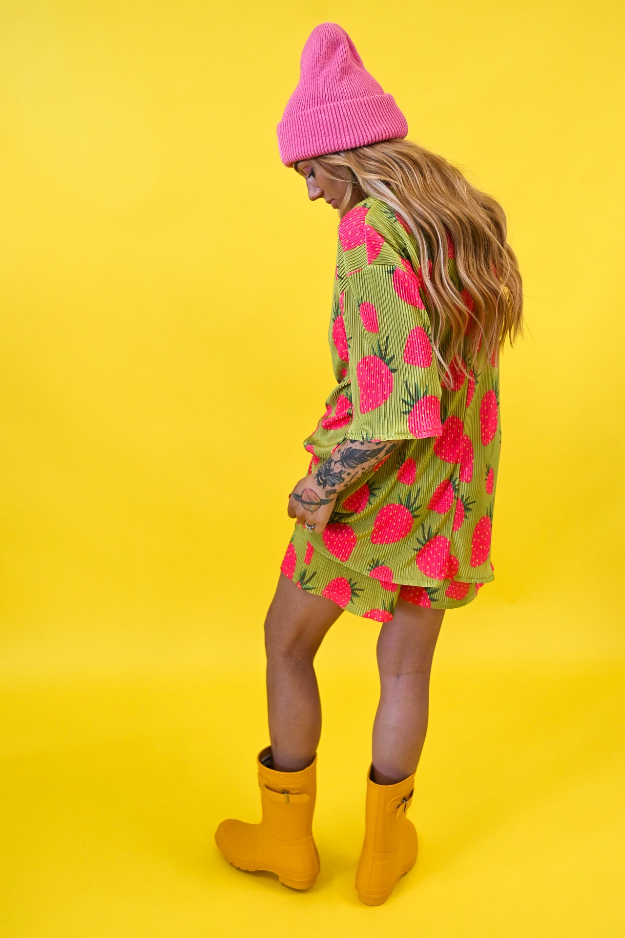 A woman wearing a pink knit beanie, a green and pink strawberry-patterned short set, and yellow rain boots, standing against a solid yellow background, back view