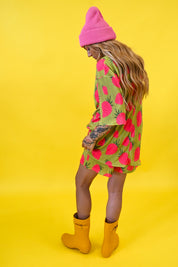 A woman wearing a pink knit beanie, a green and pink strawberry-patterned short set, and yellow rain boots, standing against a solid yellow background, back view