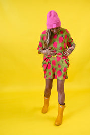 A woman modeling a short-sleeve olive green set with a bright pink and red strawberry print, styled with a pink beanie, yellow rain boots, against a solid yellow background