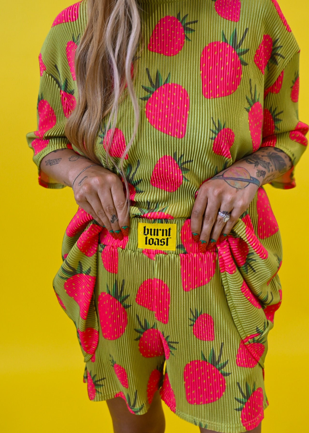 A model wearing a lime green and hot pink strawberry print plisse short set with a yellow 'BURNT TOAST' brand patch on the front, set against a solid bright yellow background holding waistband