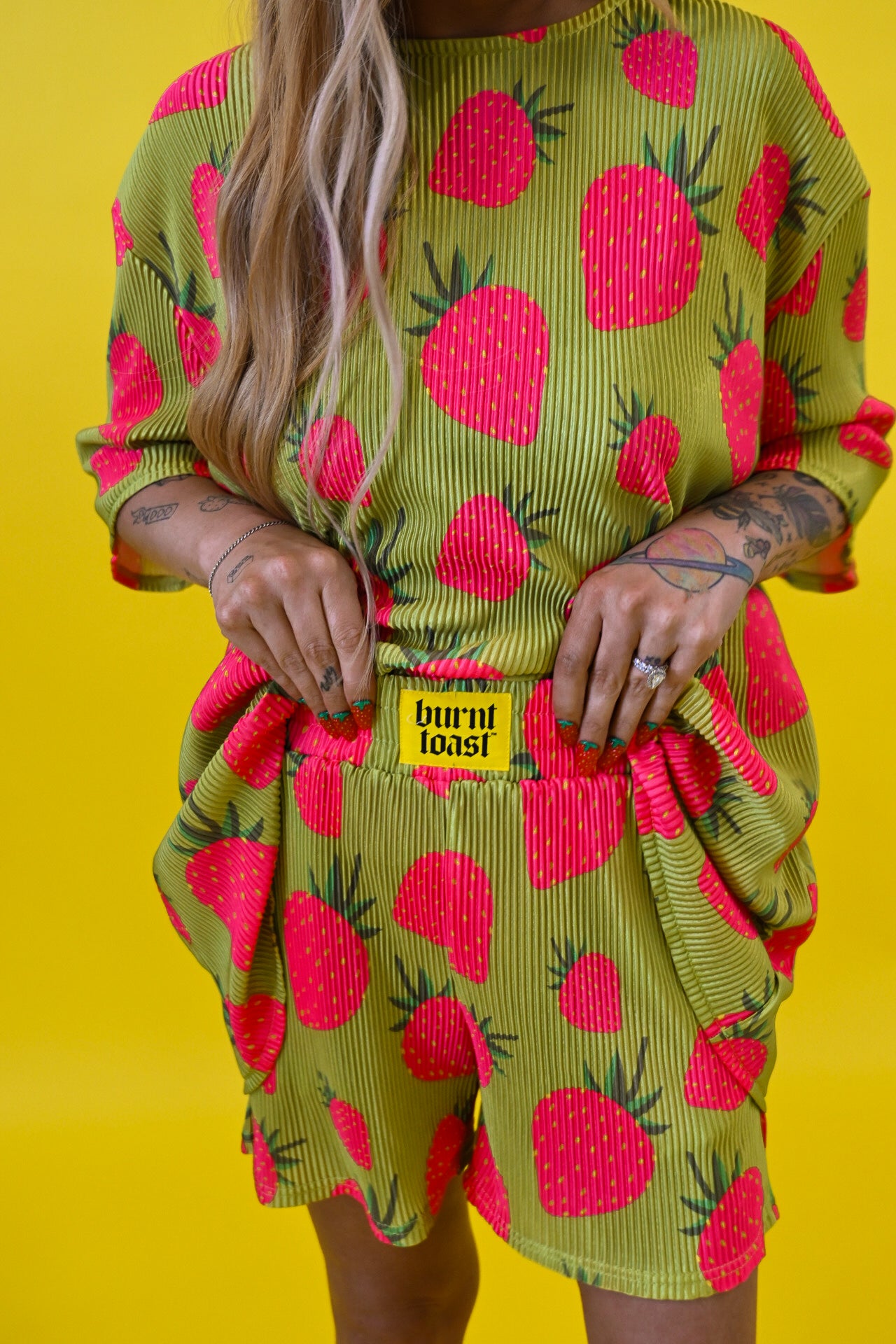 A model wearing a lime green and hot pink strawberry print plisse short set with a yellow 'BURNT TOAST' brand patch on the front, set against a solid bright yellow background holding waistband