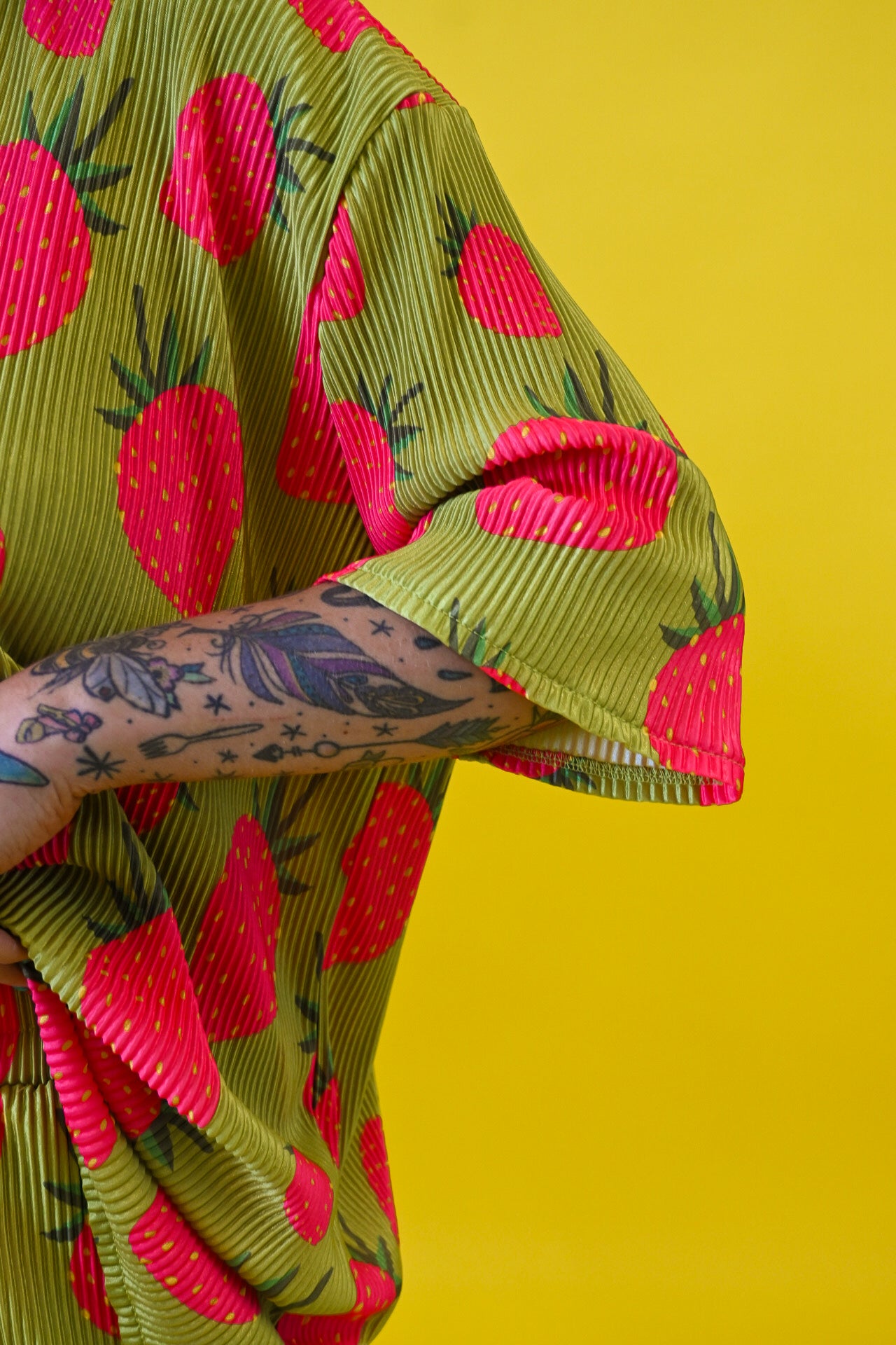 A close-up of a person wearing a short-sleeve olive green shirt with a bold pink strawberry print, set against a solid bright yellow background