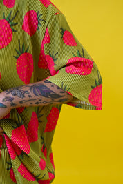 A close-up of a person wearing a short-sleeve olive green shirt with a bold pink strawberry print, set against a solid bright yellow background