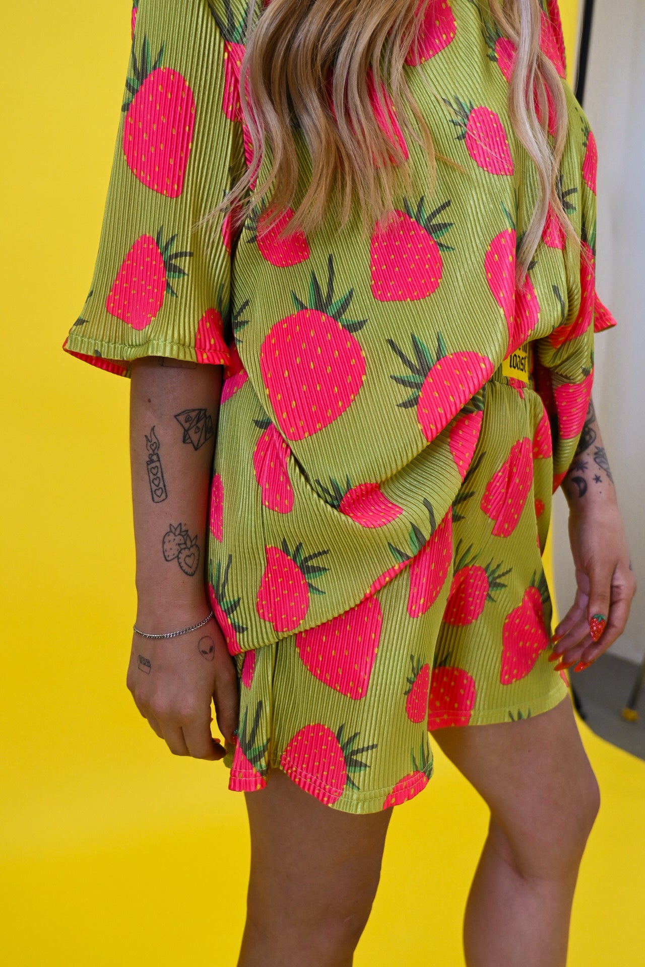 A person wearing a lime green and pink strawberry print short set, photographed from the side against a bright yellow background