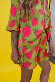 A person wearing a lime green and pink strawberry print short set, photographed from the side against a bright yellow background