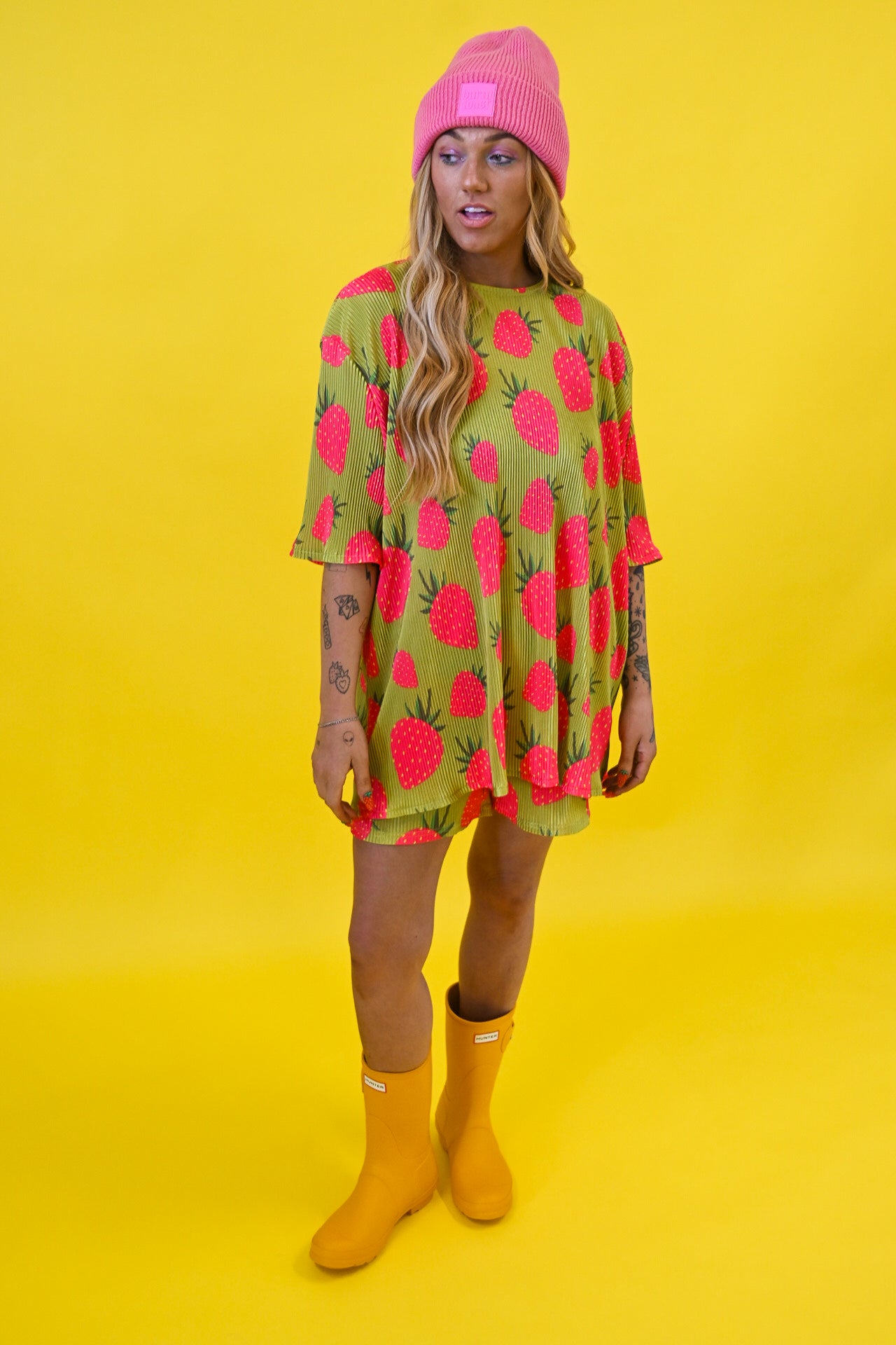 A woman modeling a short-sleeve, oversized tunic shirt with a green base and a bright pink strawberry print, paired with a pink beanie and yellow rain boots, set against a solid yellow studio background