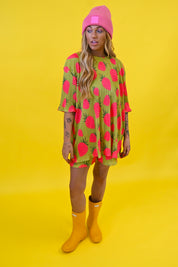 A woman modeling a short-sleeve, oversized tunic shirt with a green base and a bright pink strawberry print, paired with a pink beanie and yellow rain boots, set against a solid yellow studio background