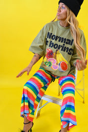 A woman wearing a black beanie, a green graphic t-shirt that reads 'Normal is boring', and rainbow-striped pants sits on a ladder against a solid bright yellow background