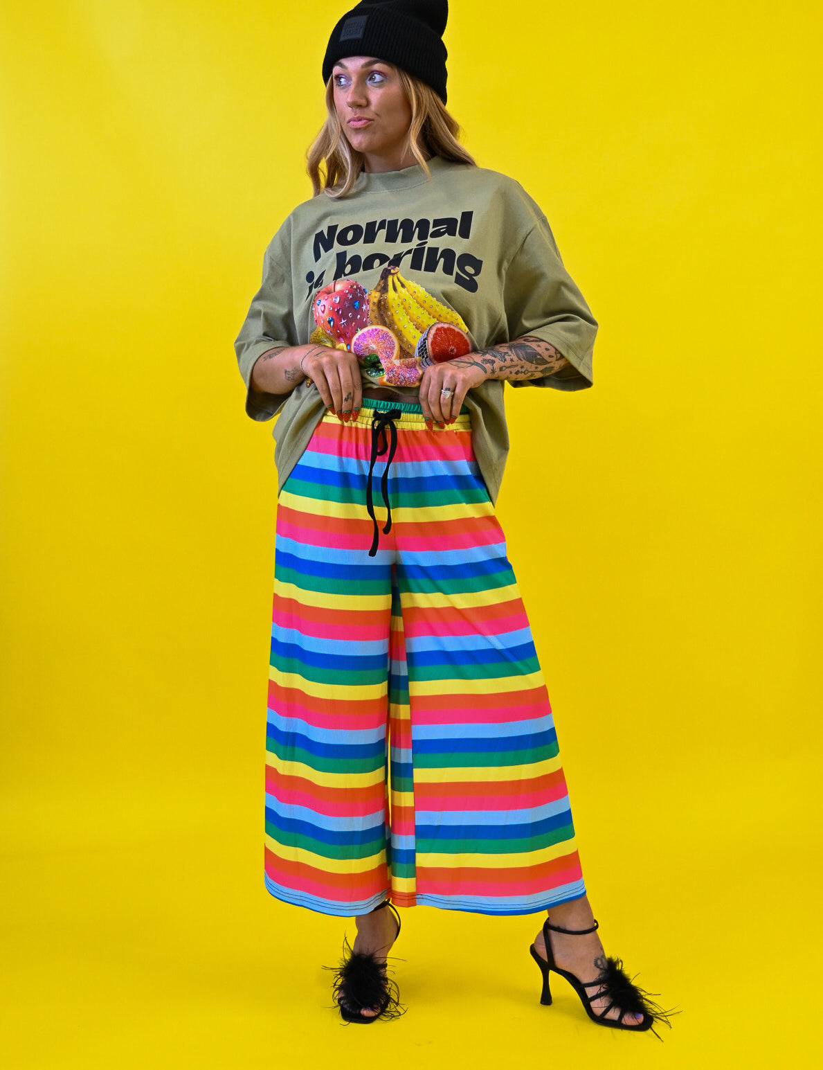 A model stands against a bright yellow studio background wearing a black beanie, a green graphic t-shirt with 'Normal is Boring' text, and wide-leg rainbow-striped pants 