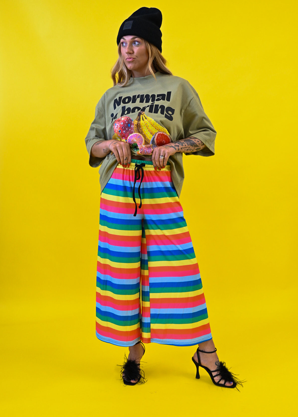 A model stands against a bright yellow studio background wearing a black beanie, a green graphic t-shirt with 'Normal is Boring' text, and wide-leg rainbow-striped pants 