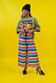 A model stands against a bright yellow studio background wearing a black beanie, a green graphic t-shirt with 'Normal is Boring' text, and wide-leg rainbow-striped pants 