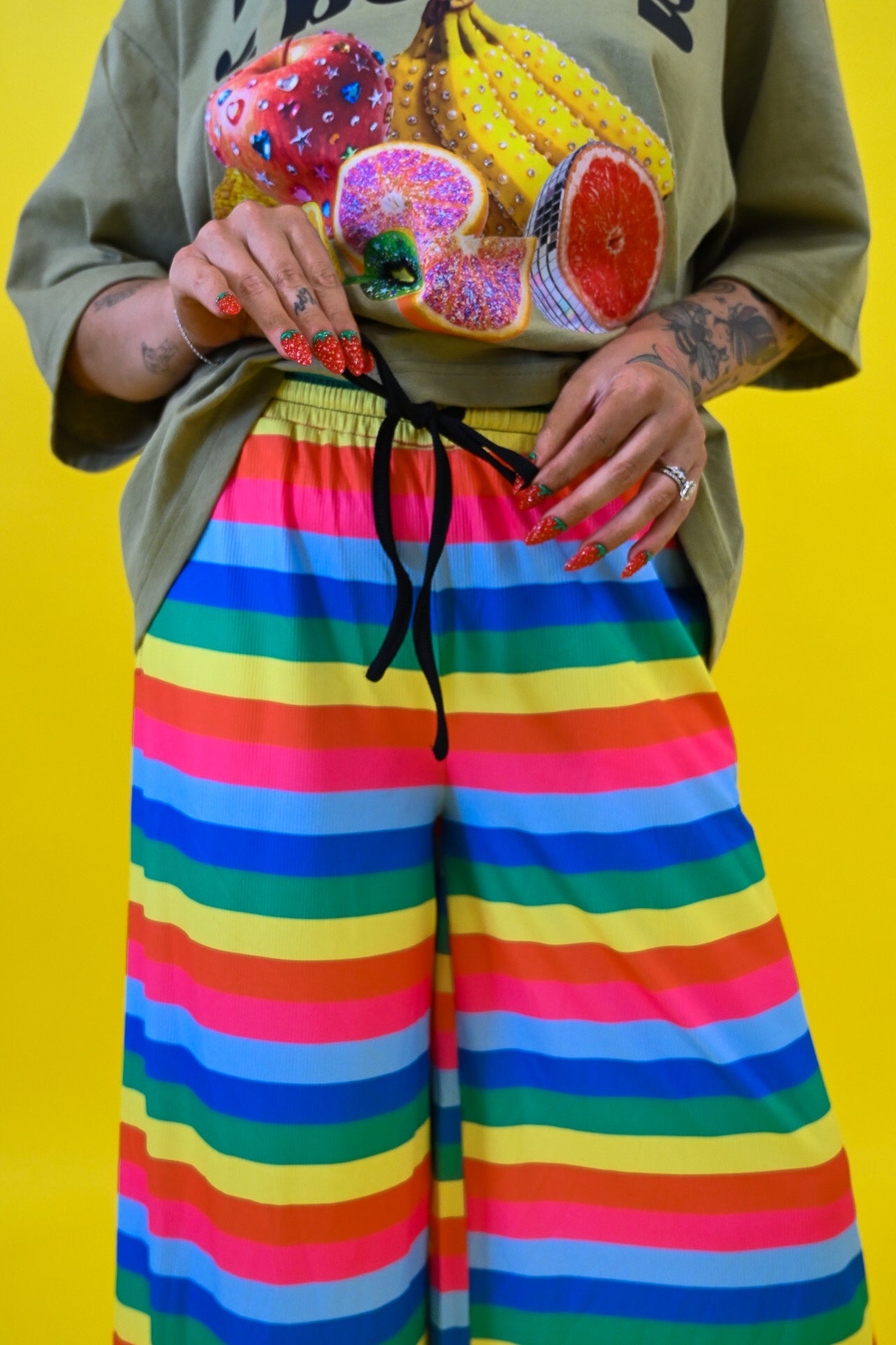 A person wearing bright rainbow-striped wide-leg pants, holding a black drawstring  at the waist, and an olive green graphic t-shirt with a fruit print, standing against a solid yellow background