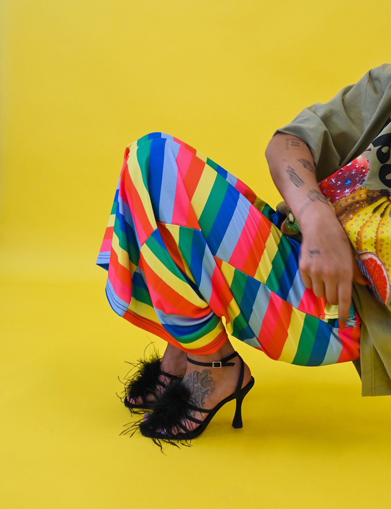 A person seated against a bright yellow studio background, wearing loose multicolored striped pants, an olive green top, and black high-heeled sandals with feather trim
