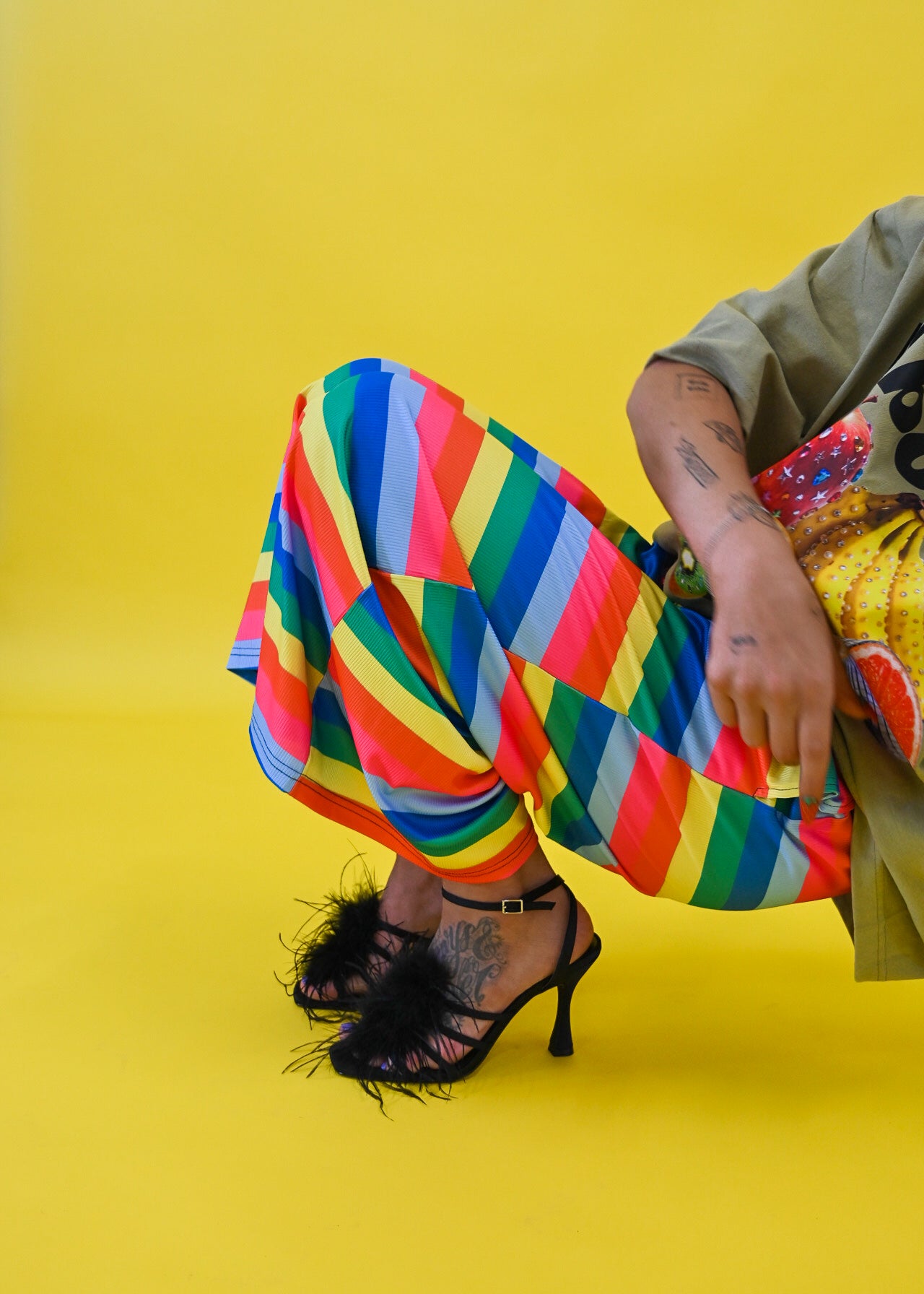 A person seated against a bright yellow studio background, wearing loose multicolored striped pants, an olive green top, and black high-heeled sandals with feather trim