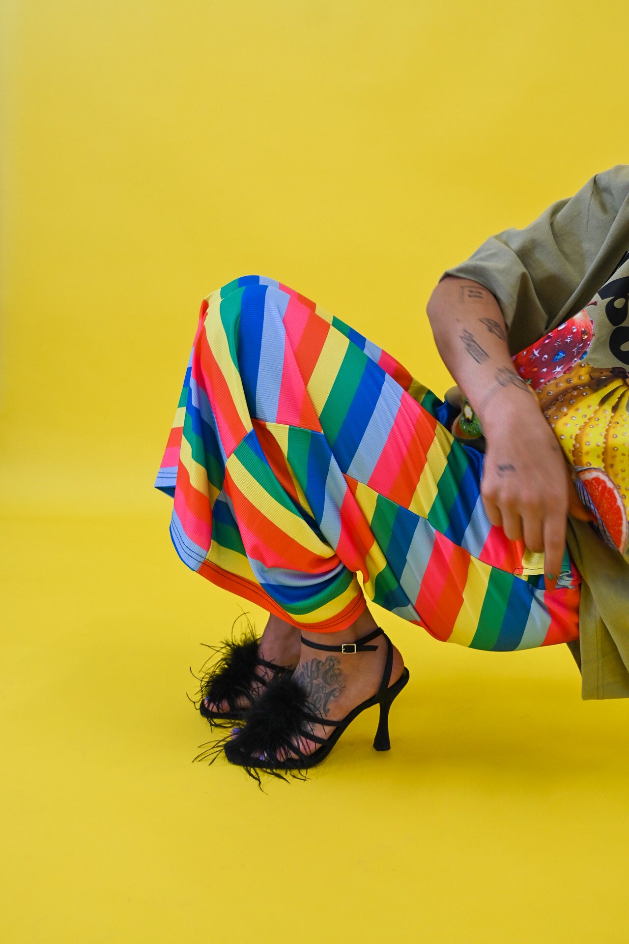 A person seated against a bright yellow studio background, wearing loose multicolored striped pants, an olive green top, and black high-heeled sandals with feather trim