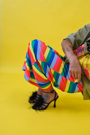 A person seated against a bright yellow studio background, wearing loose multicolored striped pants, an olive green top, and black high-heeled sandals with feather trim