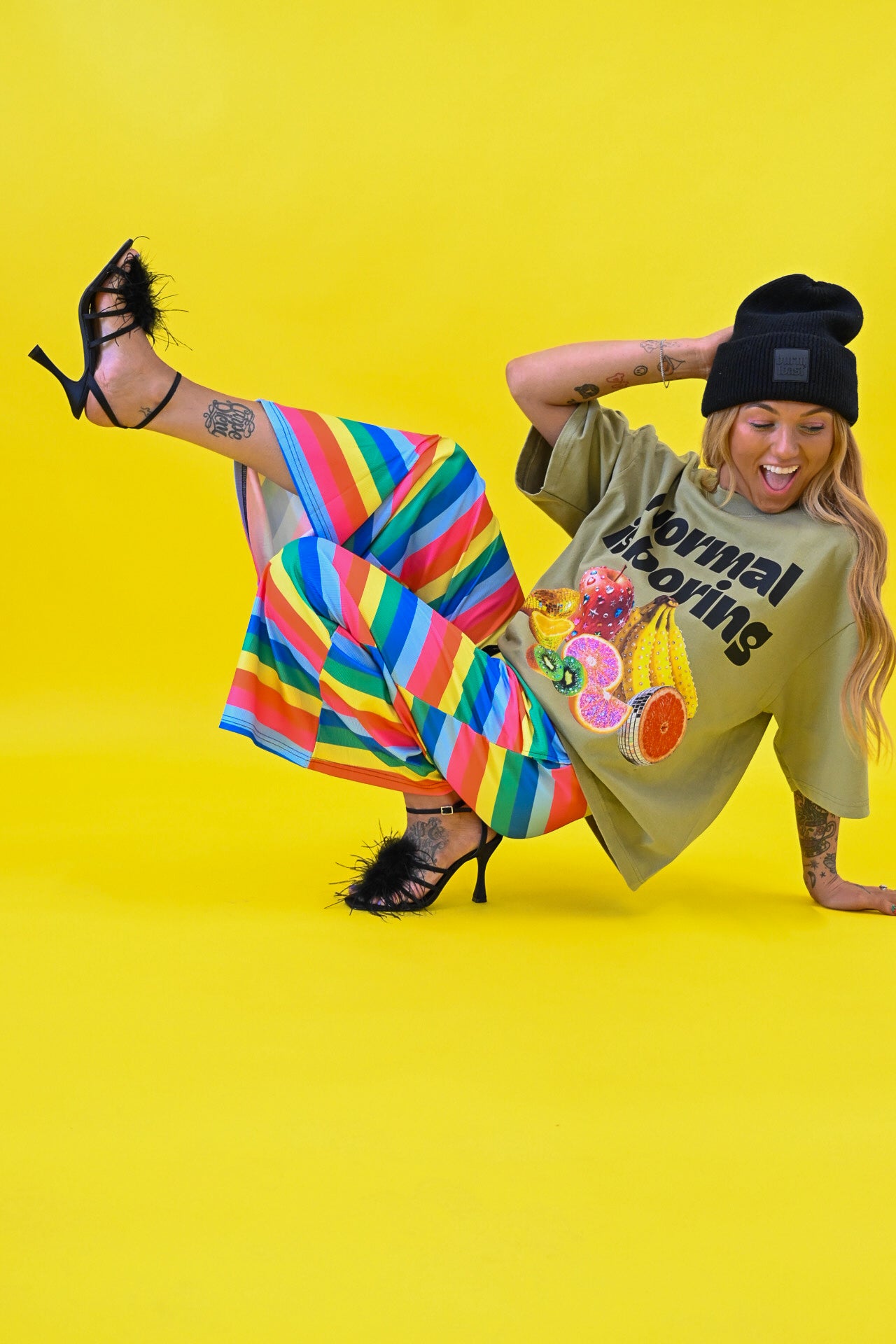 A woman poses on a bright yellow studio background wearing a black beanie, black strappy heels, a beige graphic t-shirt, and vibrant rainbow-striped pants