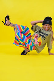 A woman poses on a bright yellow studio background wearing a black beanie, black strappy heels, a beige graphic t-shirt, and vibrant rainbow-striped pants