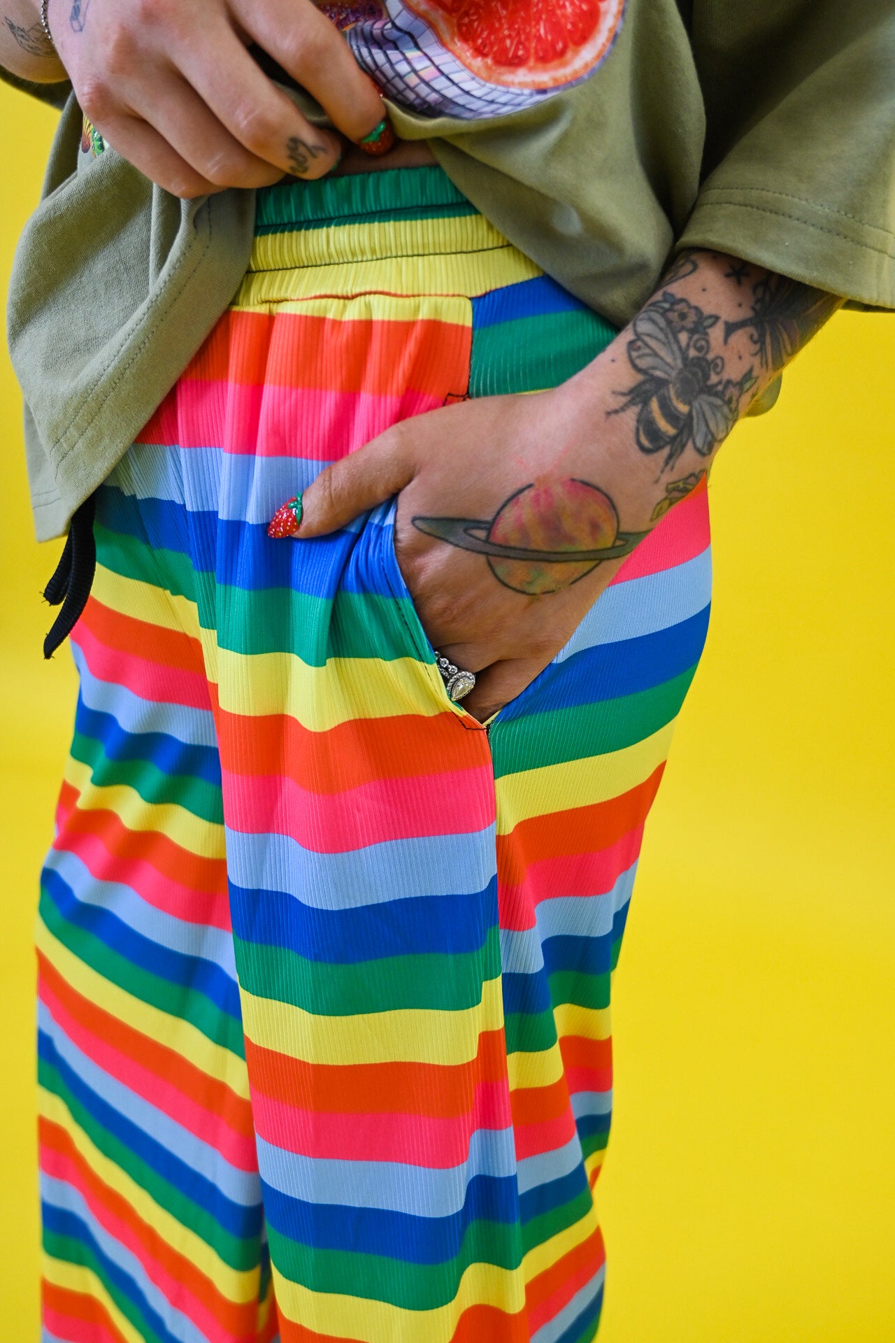 A close-up of a person wearing bright rainbow-striped pants with a side pocket, holding a green shirt open, against a solid yellow background