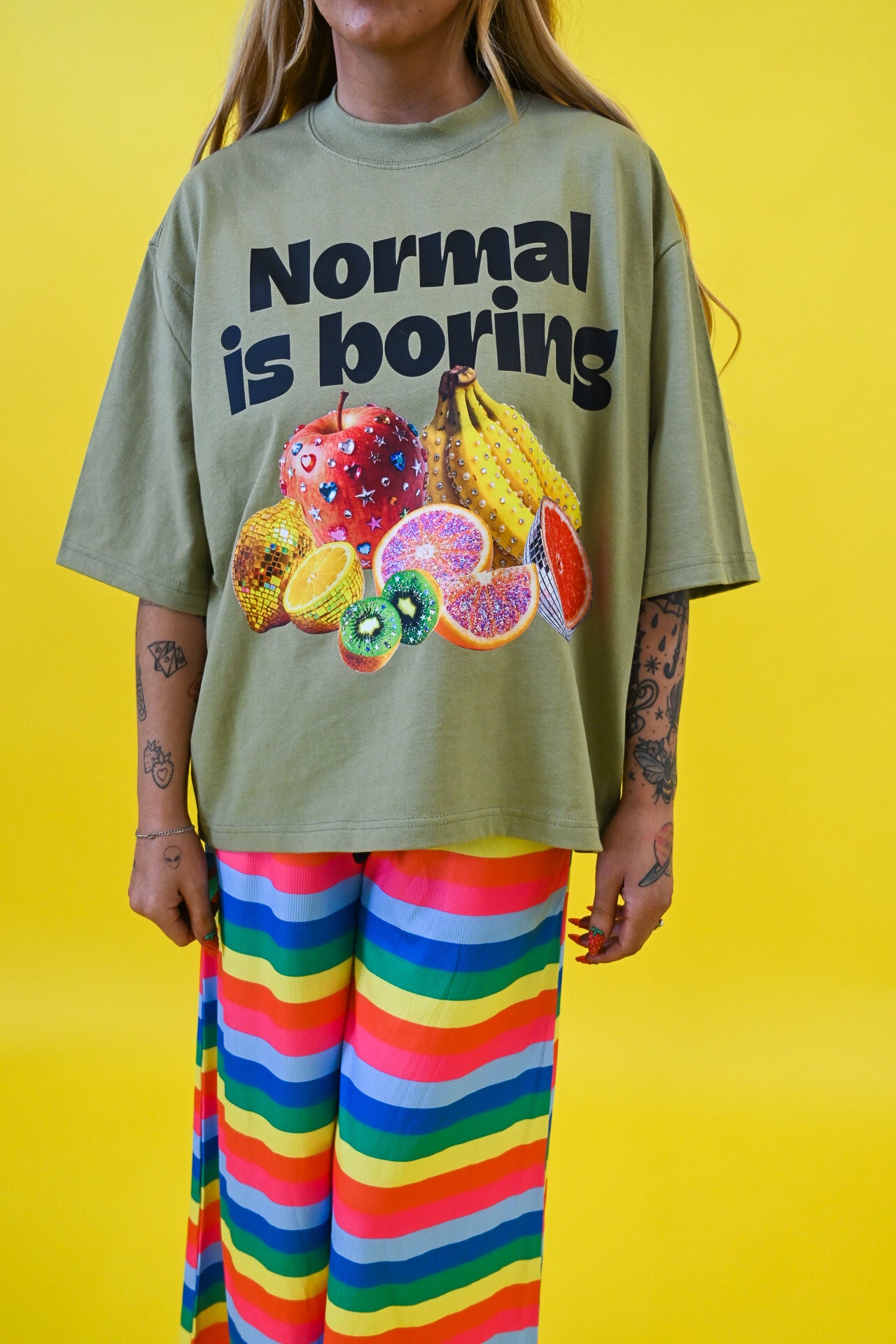 A person wearing an oversized sage green graphic T-shirt with the text 'Normal is boring' and a colorful fruit illustration, paired with bright rainbow-striped pants, against a solid yellow background