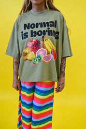 A person wearing an oversized sage green graphic T-shirt with the text 'Normal is boring' and a colorful fruit illustration, paired with bright rainbow-striped pants, against a solid yellow background