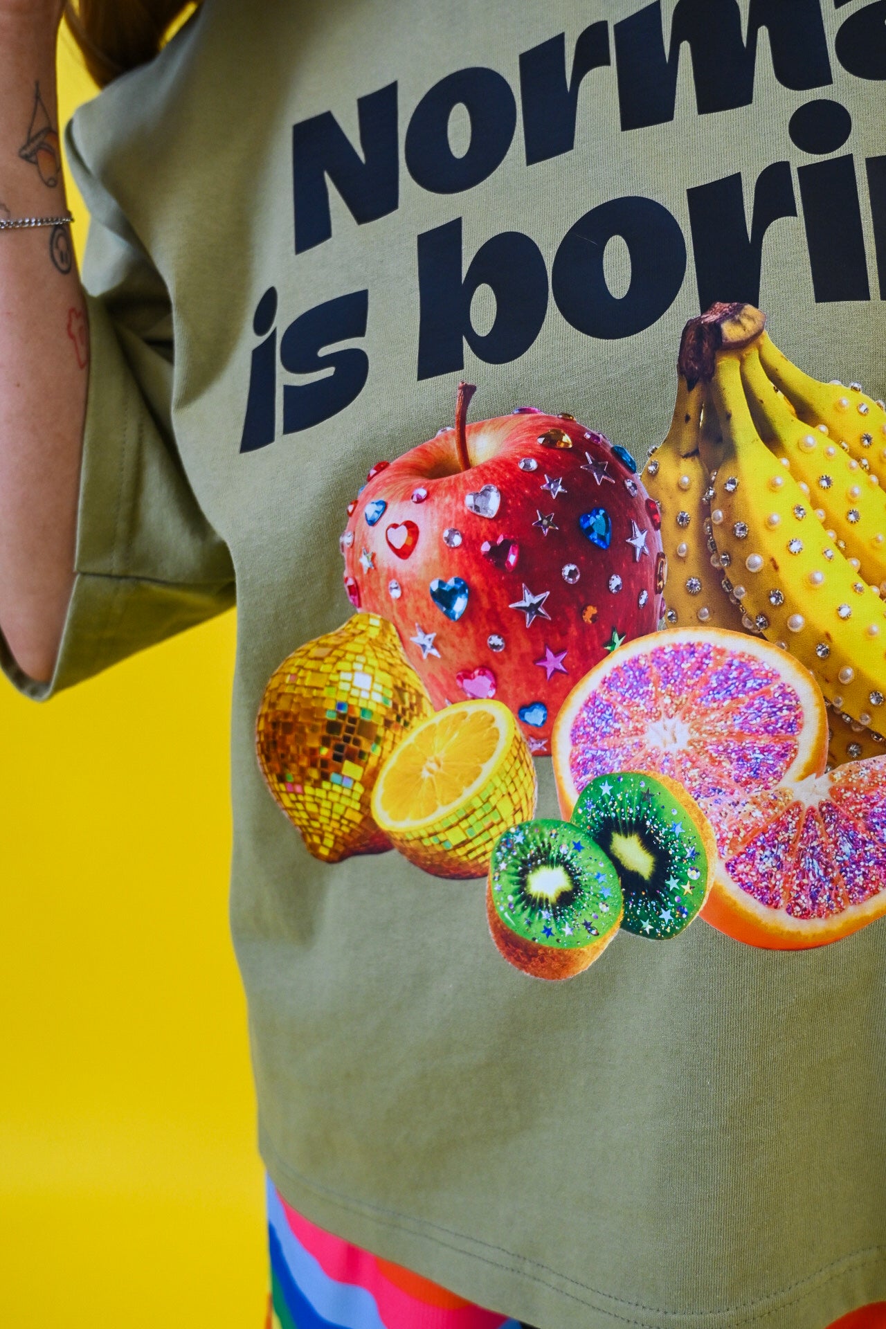 A close-up of a person wearing a light olive green graphic T-shirt with the text 'Normal is boring' in bold black letters, featuring a colorful fruit print including a red apple, bananas, orange, kiwi, and blood orange. The background is a solid bright yellow