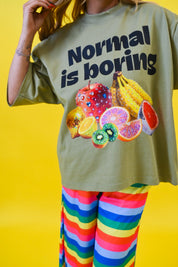 A person wearing an oversized olive green graphic T-shirt with the text 'Normal is boring' and a colorful fruit print, paired with bright rainbow-striped pants, against a solid yellow background