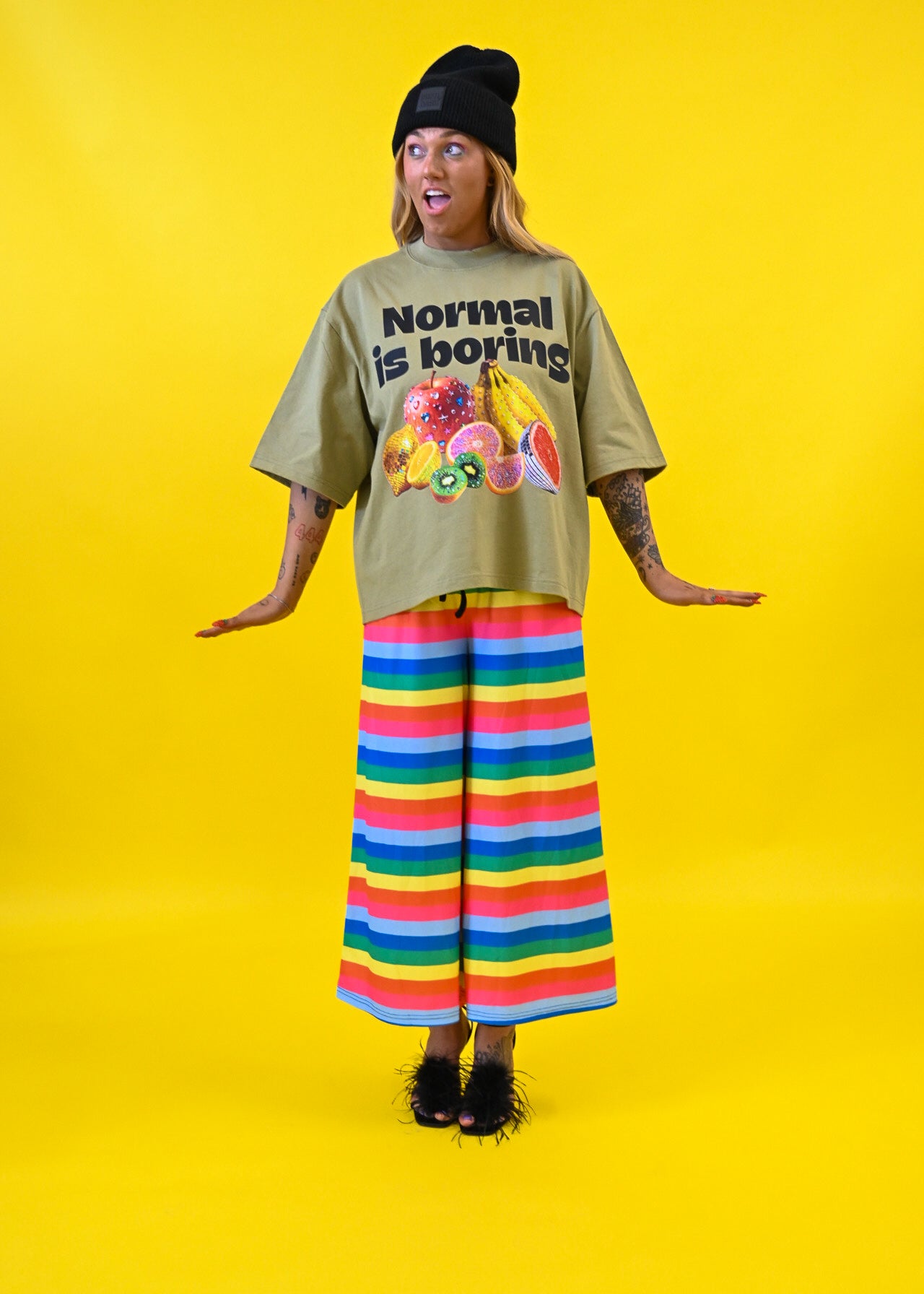 A model stands against a bright yellow studio background wearing an oversized olive green graphic t-shirt with the text 'Normal is boring' and a fruit illustration, paired with wide-leg pants featuring horizontal rainbow stripes, a black beanie, and black shoes