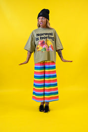 A model stands against a bright yellow studio background wearing an oversized olive green graphic t-shirt with the text 'Normal is boring' and a fruit illustration, paired with wide-leg pants featuring horizontal rainbow stripes, a black beanie, and black shoes