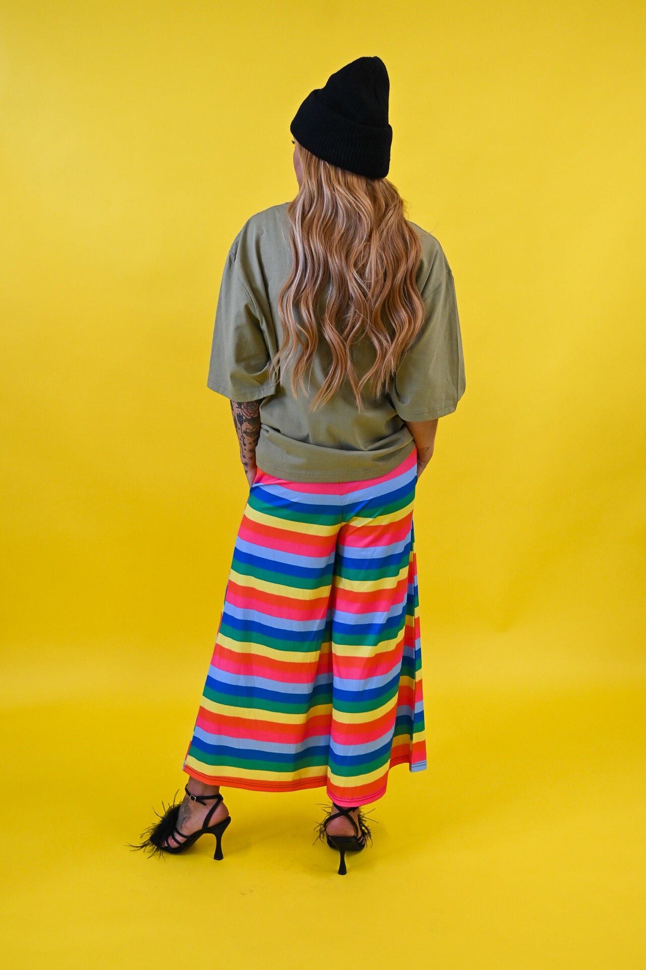 A person shown from the back wearing a black beanie, olive green t-shirt, and a colorful rainbow-striped midi pants, standing against a solid bright yellow background