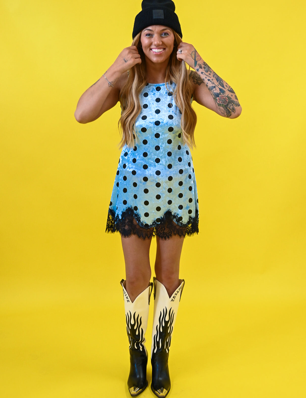 A woman modeling a light blue sleeveless dress with black polka dots and a black lace hem, styled with a black beanie, cowboy boots, and arm tattoos against a solid yellow background