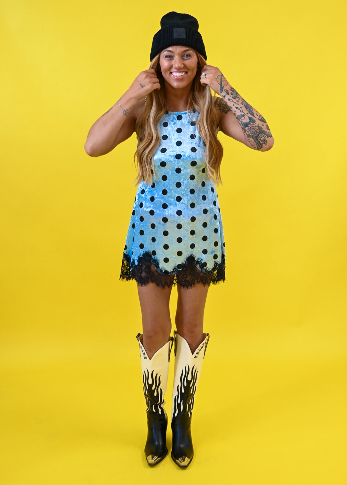 A woman modeling a light blue sleeveless dress with black polka dots and a black lace hem, styled with a black beanie, cowboy boots, and arm tattoos against a solid yellow background