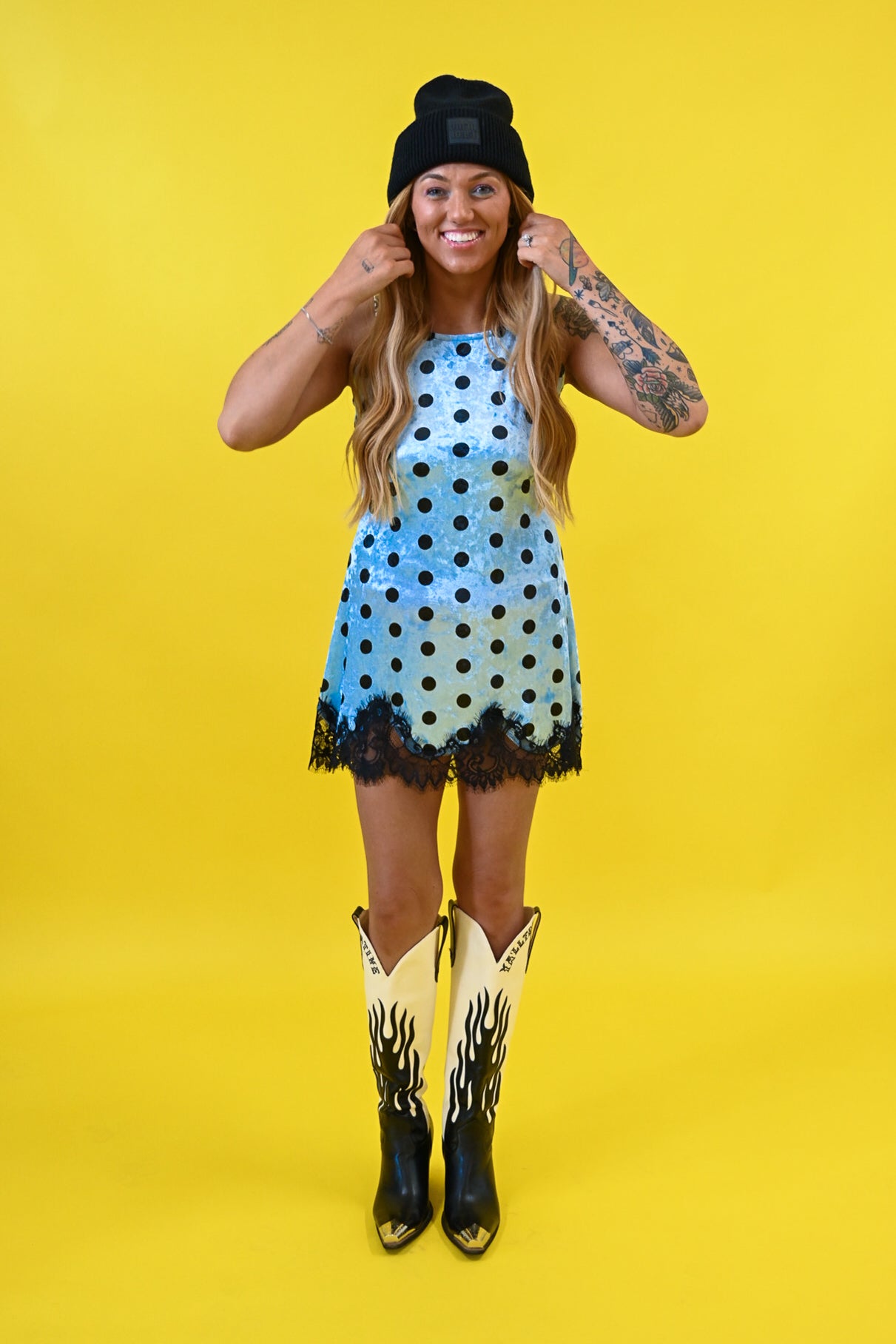 A woman modeling a light blue sleeveless dress with black polka dots and a black lace hem, styled with a black beanie, cowboy boots, and arm tattoos against a solid yellow background