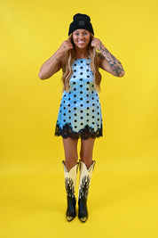 A woman modeling a light blue sleeveless dress with black polka dots and a black lace hem, styled with a black beanie, cowboy boots, and arm tattoos against a solid yellow background