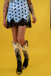 A model wearing a light blue and black polka dot mini dress with a black lace hem, paired with black and cream cowboy boots featuring flame designs, set against a solid yellow background, close up view