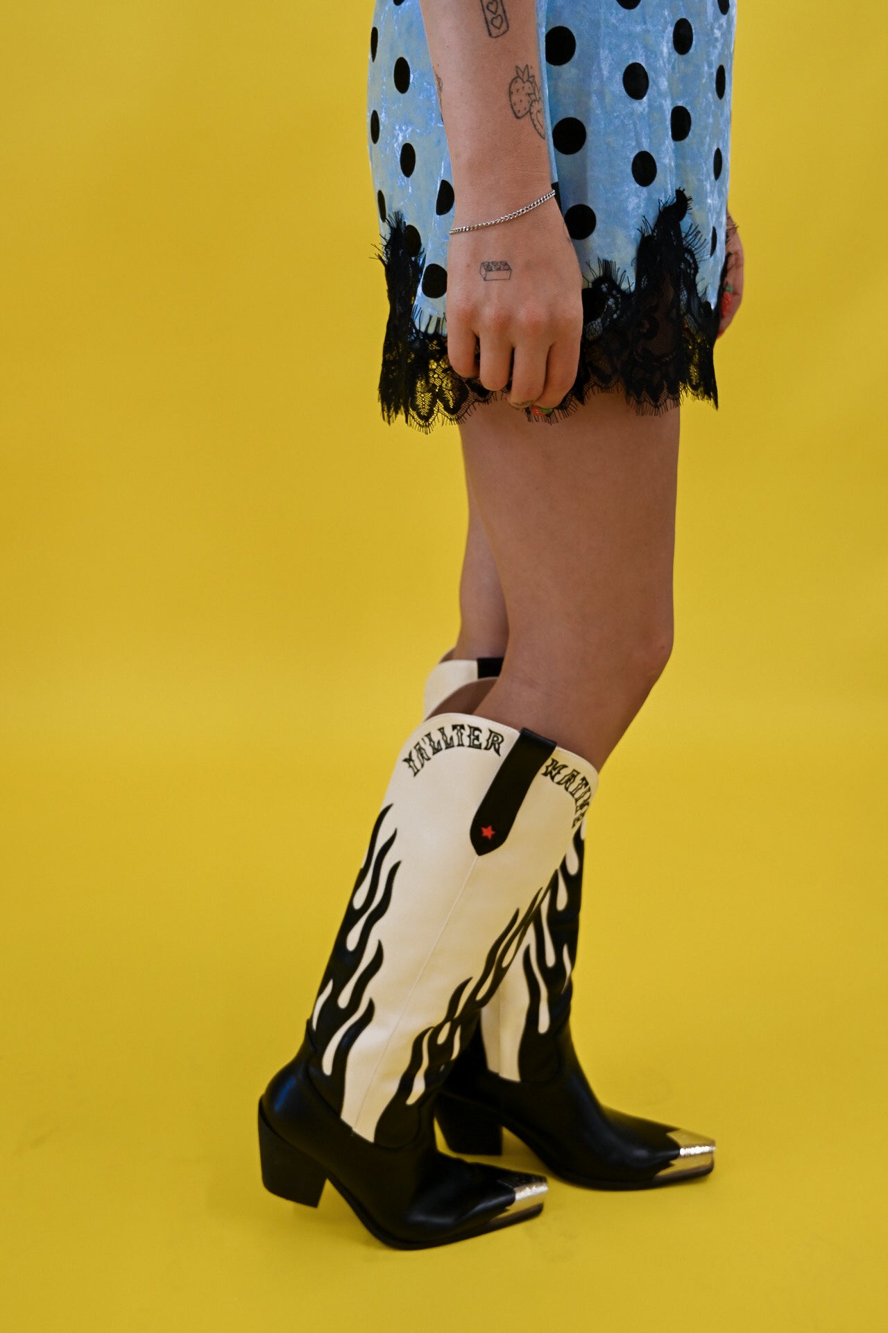 A person wearing black and white western-style heeled boots with flame-like white patterns and silver toe caps, standing against a bright yellow studio background. The person is also wearing a light blue polka-dot skirt with black lace trim