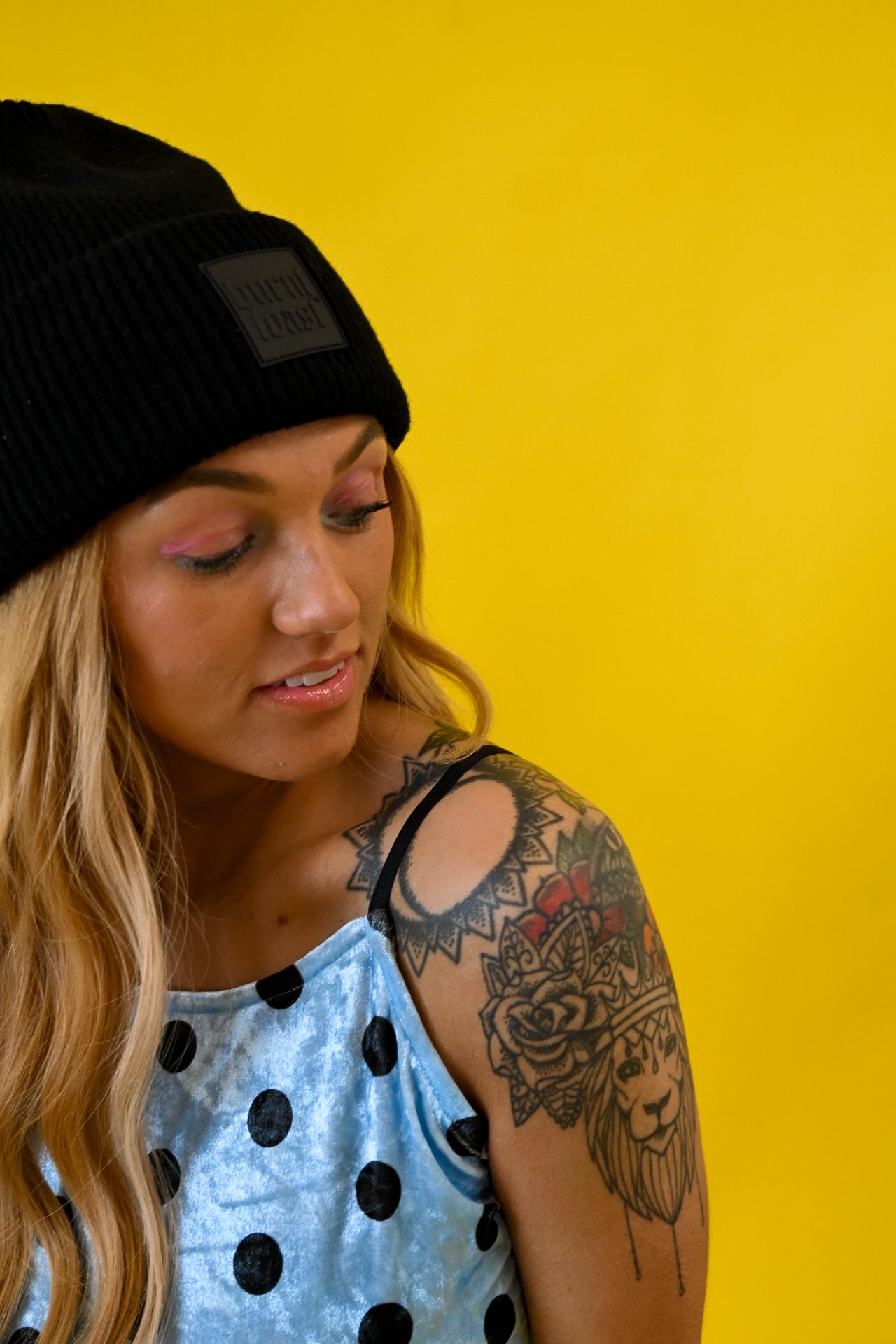 A woman with blonde hair, a black beanie, and a blue polka-dot top, looking down against a solid yellow background. She has a large floral tattoo on her upper arm