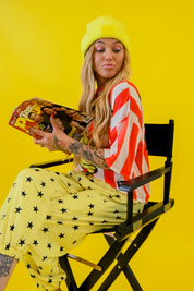 A woman sitting in a black director's chair against a solid yellow background, wearing a bright yellow beanie, a red and white striped long-sleeve jersey, and yellow pants with black star prints. She is holding a magazine