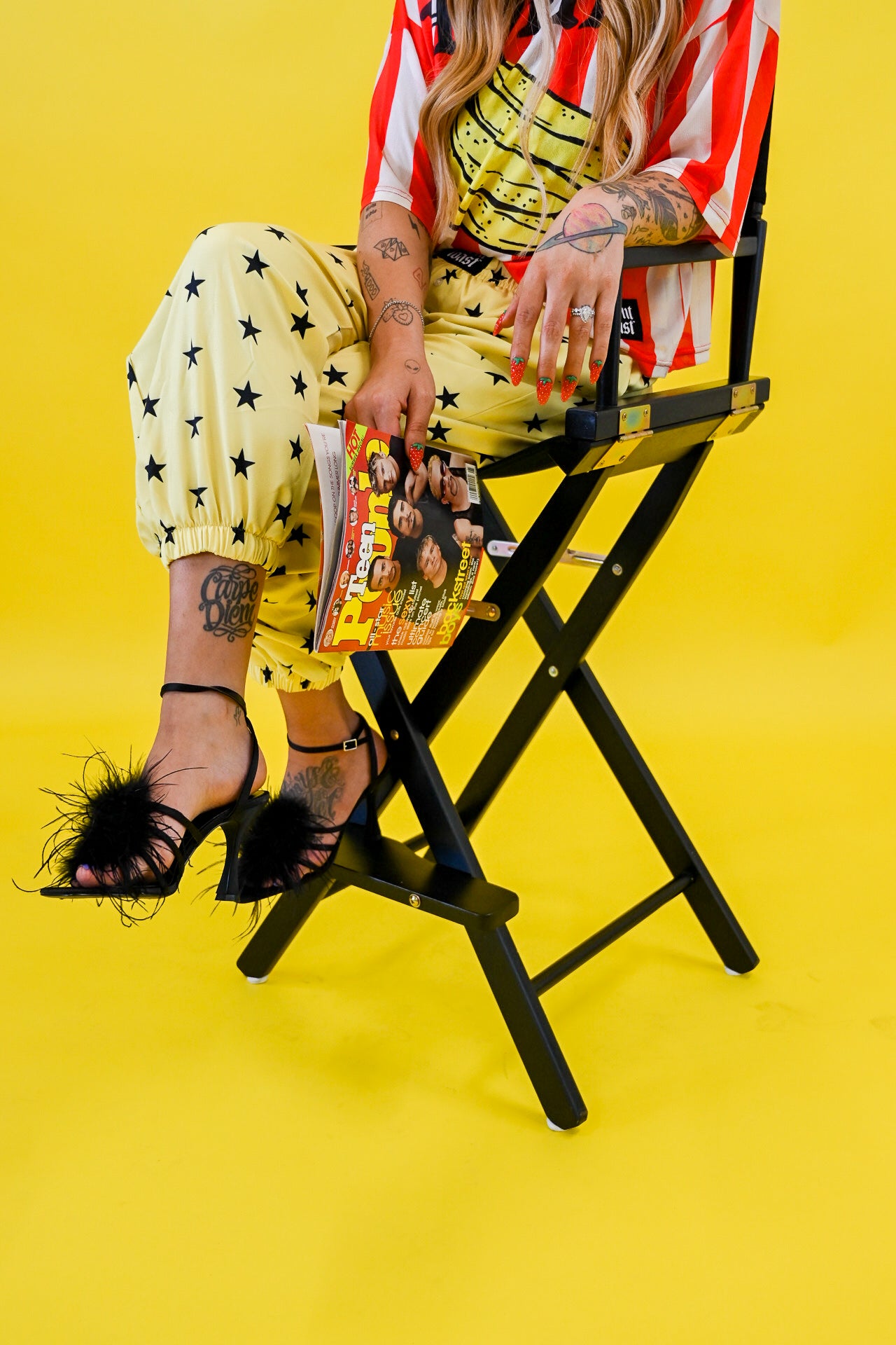 A model sits on a black folding director's chair against a bright yellow studio background, wearing a red and white striped jersey, yellow pants with black star prints, and black feather-embellished high heels. She holds a colorful magazine
