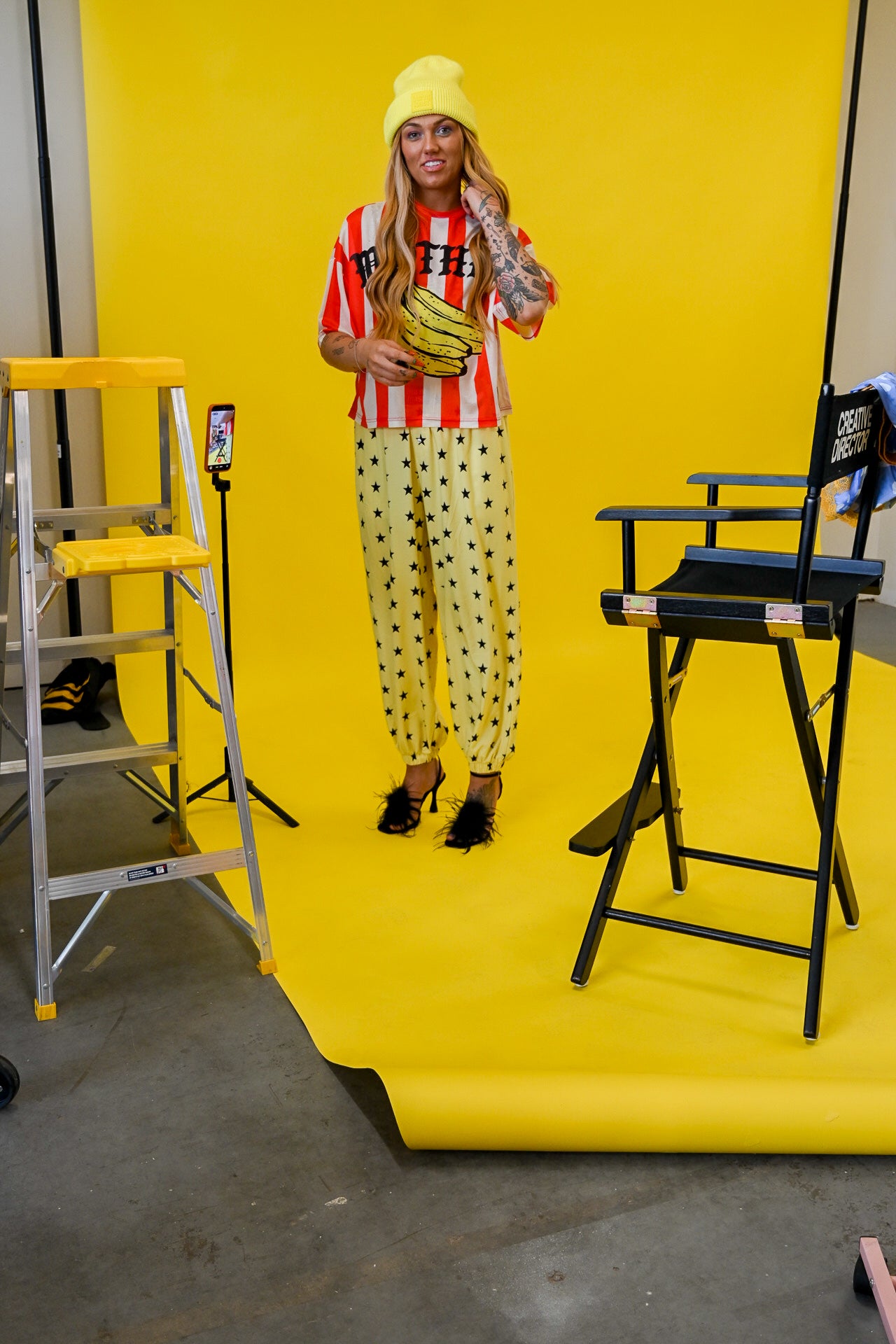 A woman stands in a studio wearing a bright yellow outfit with red-and-white striped sleeves, yellow polka-dot pants, and a yellow beanie, while posing in front of a yellow backdrop. A ladder, chair, and phone stand are visible around her
