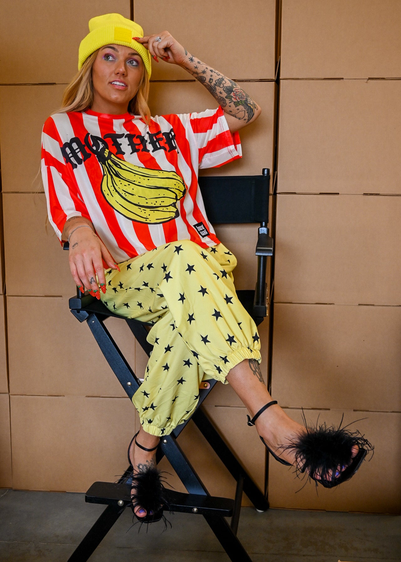 A woman sits in a black director's chair wearing a red and white striped t-shirt with a banana graphic and the word 'MOTHER', paired with yellow star-print pants, a yellow beanie, and black feather-embellished sandals against a tan paneled wall