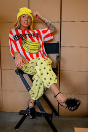 A woman sits in a black director's chair wearing a red and white striped t-shirt with a banana graphic and the word 'MOTHER', paired with yellow star-print pants, a yellow beanie, and black feather-embellished sandals against a tan paneled wall
