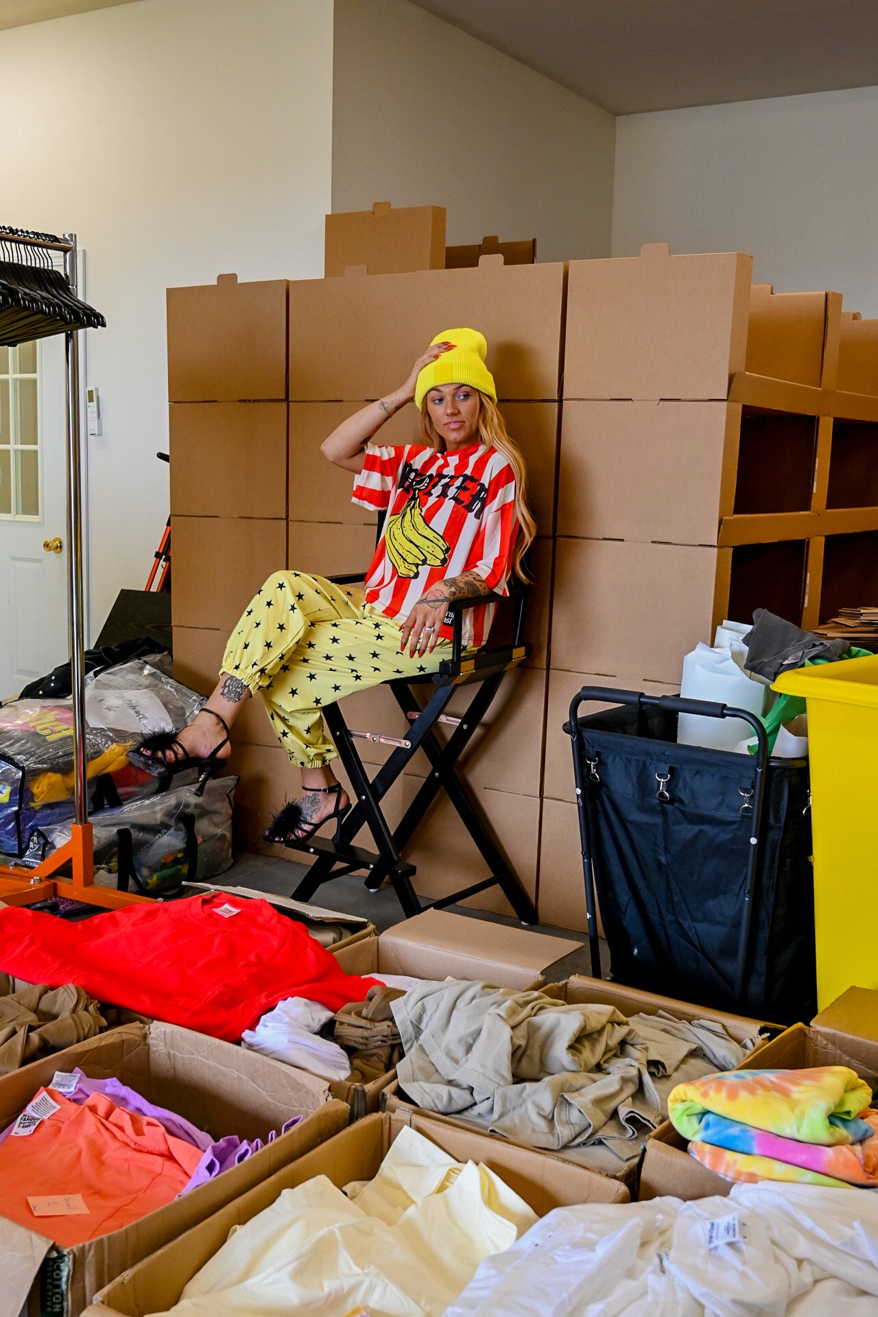 A person sits in a black folding chair in a cluttered room, wearing a red and white striped banana-themed jersey, yellow polka-dot pants, and a yellow beanie. Behind them are stacked brown cardboard boxes, with open boxes in the foreground containing folded clothing