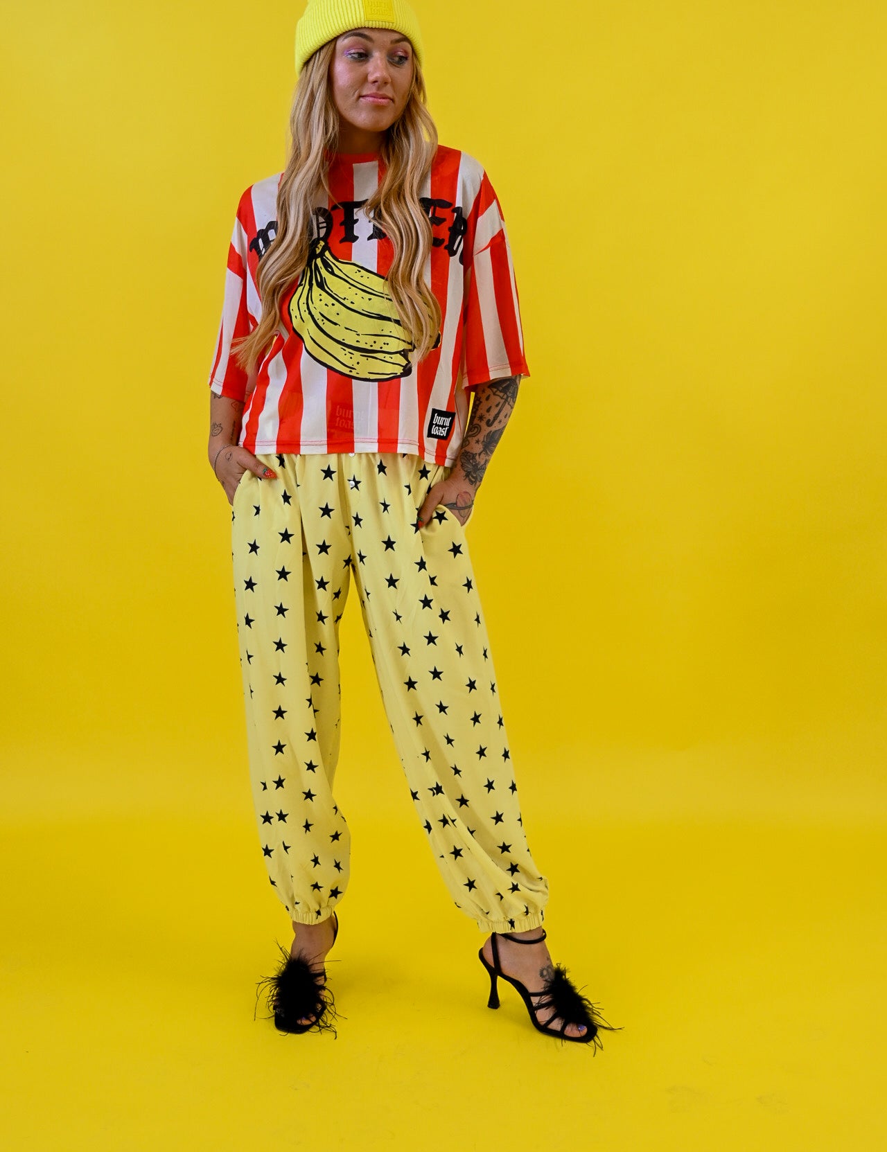 A model stands against a solid bright yellow studio background wearing a yellow beanie, a red and white striped graphic mesh jersey with a banana print, yellow jogger pants with black star patterns, and black strappy heels